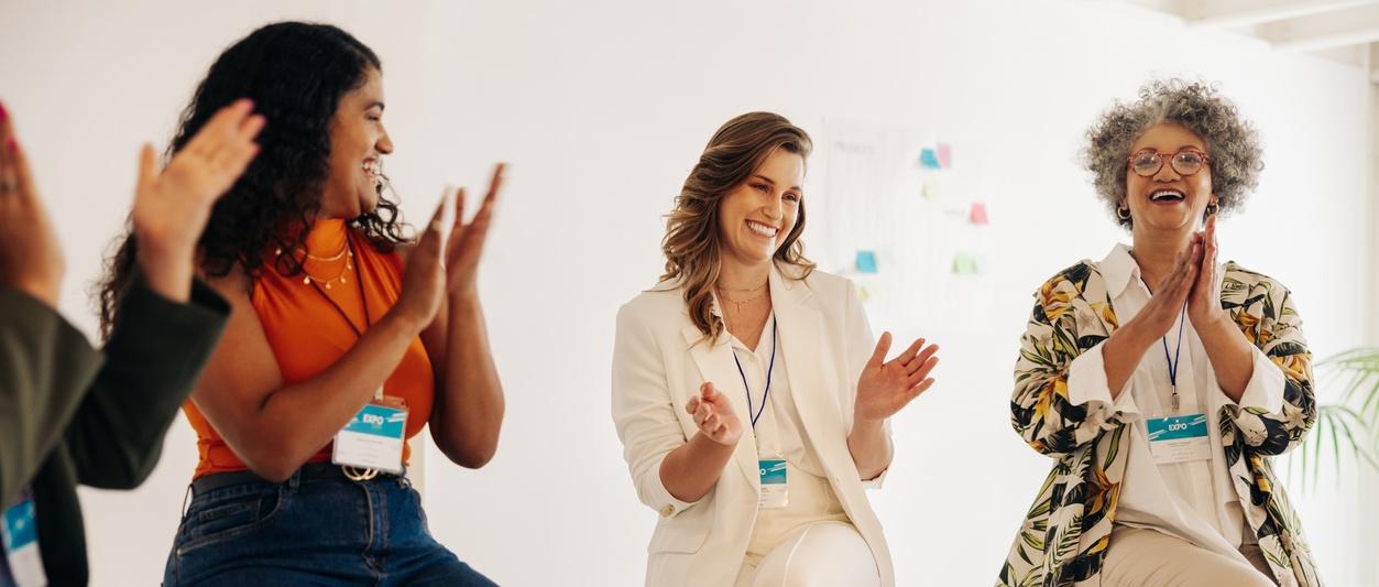 Photo of three women wearing employee lanyards clapping in a meeting