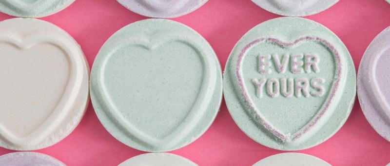 Green, purple and peach pastel-coloured Love Hearts sweets against a pink background. One Love Heart reads: EVER YOURS