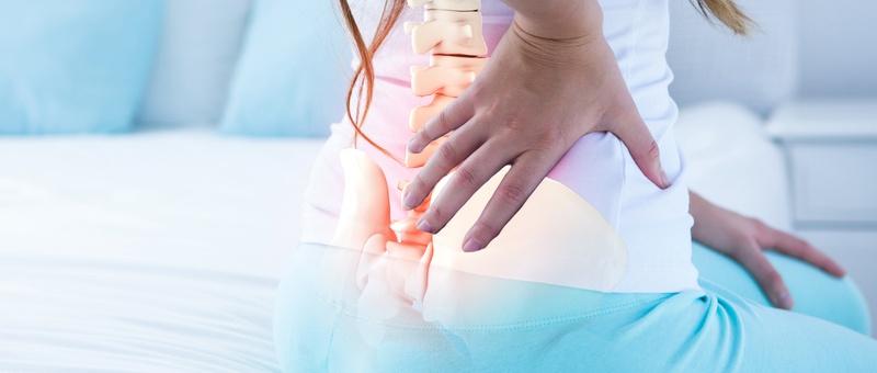 Female figure holding back on bed against pale minimalist background. Overlapping graphic of spine and pelvis.