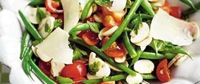 Fresh green bean salad with tomatoes, cheese shavings and herbs served in a white scalloped bowl on checkered cloth.