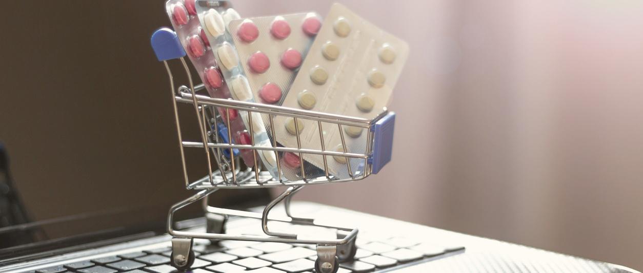 Miniature shopping trolley filled with blister packs of pink and white birth control tablets, sitting on a laptop keyboard.