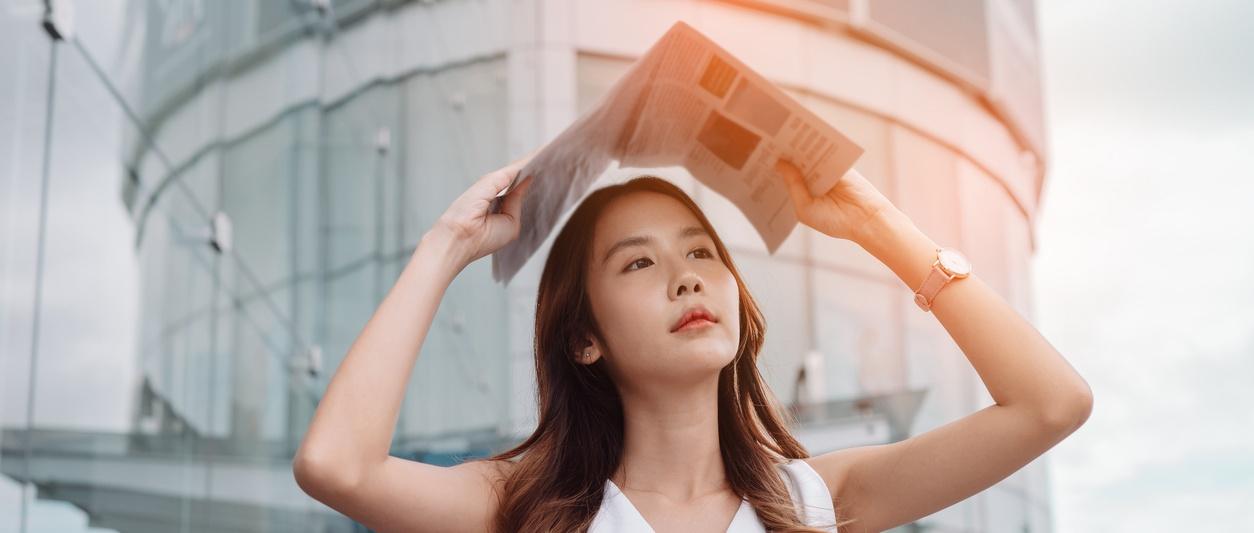 Person holding newspaper above head outside modern glass building with warm sunlight creating orange glow.