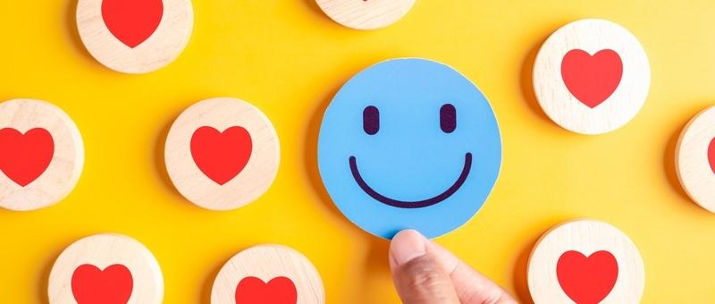 Pattern of red hearts printed on pale wooden circles with hand holding large blue emoji-style smiley face against bright yellow background.