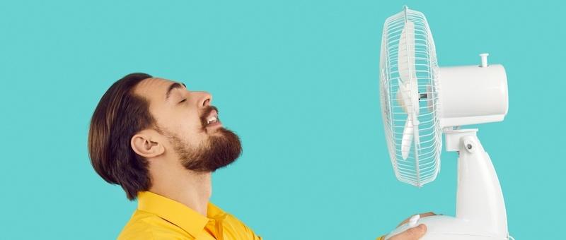 Funny bearded man enjoying breeze from electric fan against a blue background.