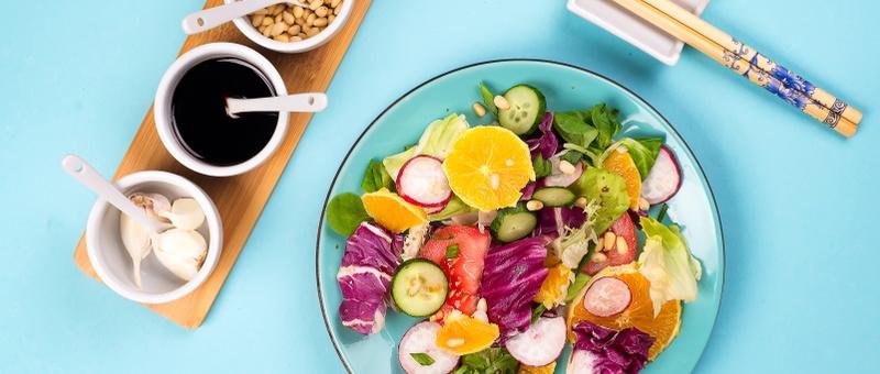White plate filled with fresh vegetarian salad alongside chopsticks, garlic, nuts and sauce selection, against a blue background.