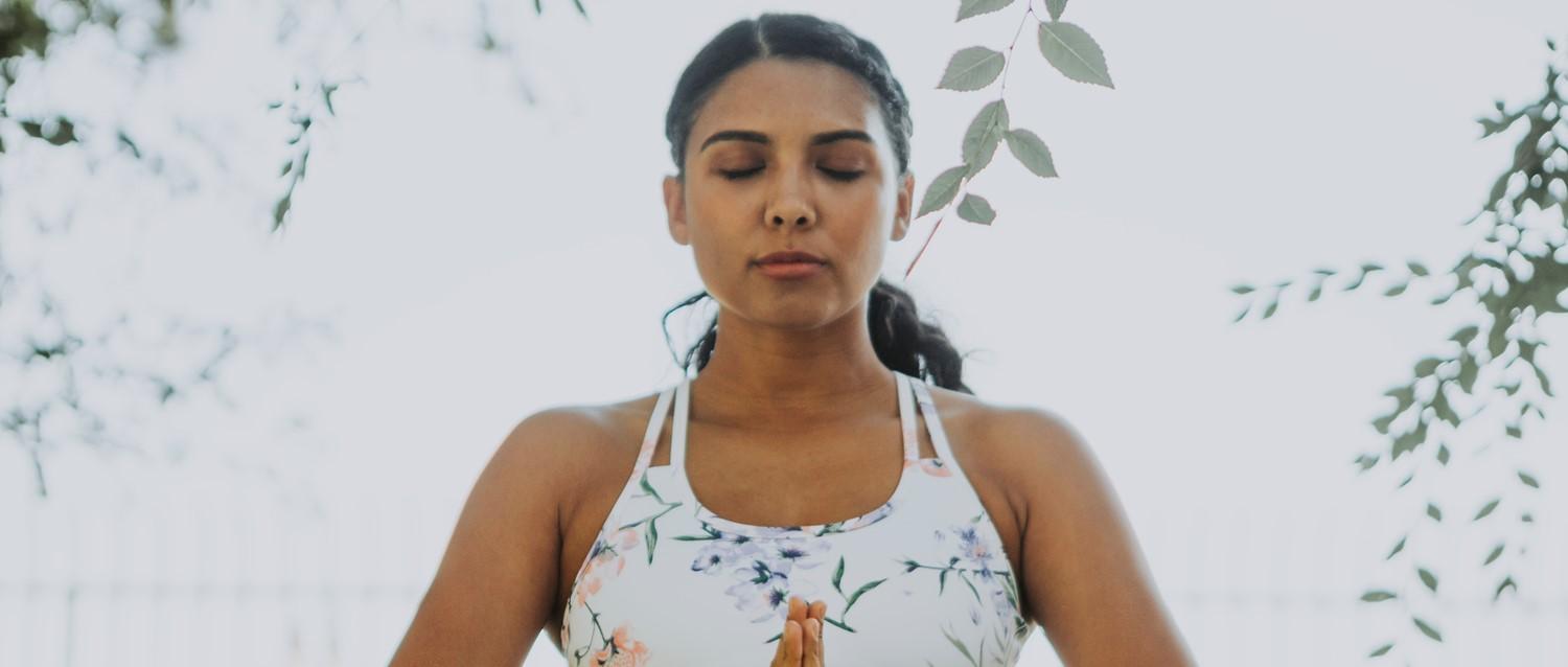 Person in floral yoga top meditating with closed eyes, surrounded by delicate green foliage in a bright setting.
