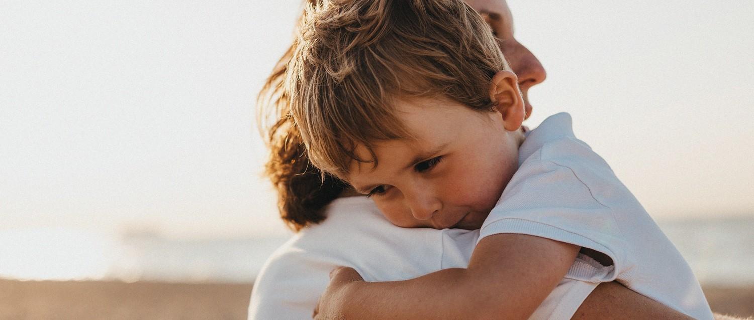 Young child with brown hair in white shirt embracing adult during golden hour outdoors.
