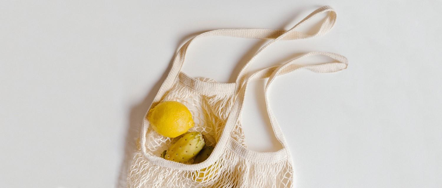 A cream-coloured reusable mesh shopping bag containing a bright yellow lemon and bananas on a white background.