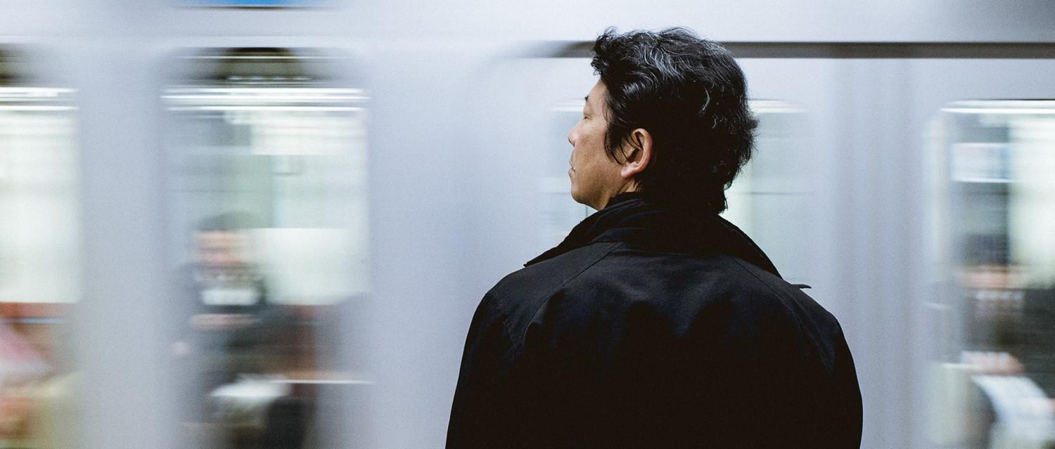 Person in black coat waiting at subway platform with blurred train passing in background.