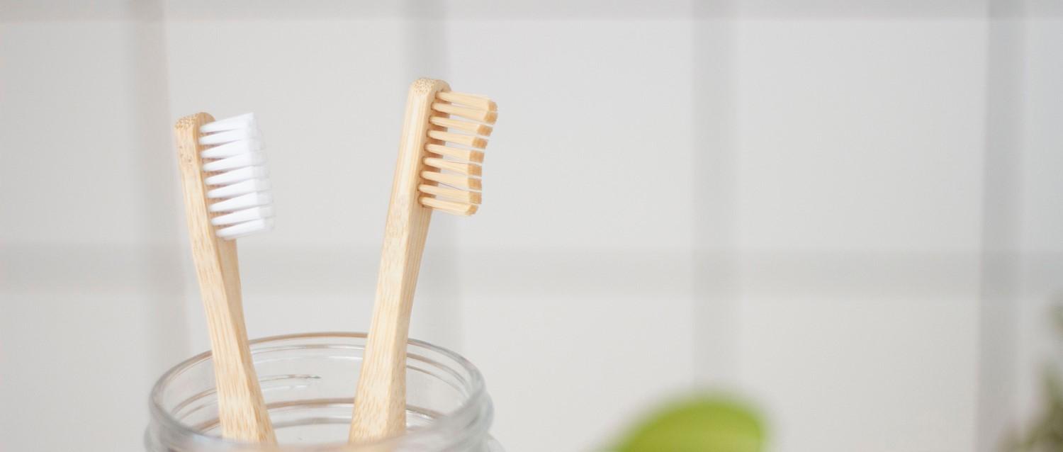 Two bamboo toothbrushes in a glass jar against a white background, one with white bristles and one with natural bristles.