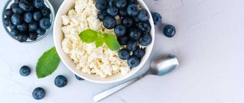 Bowl of cottage cheese and blueberries beside metal spoon and mint leaves against pale purple background.